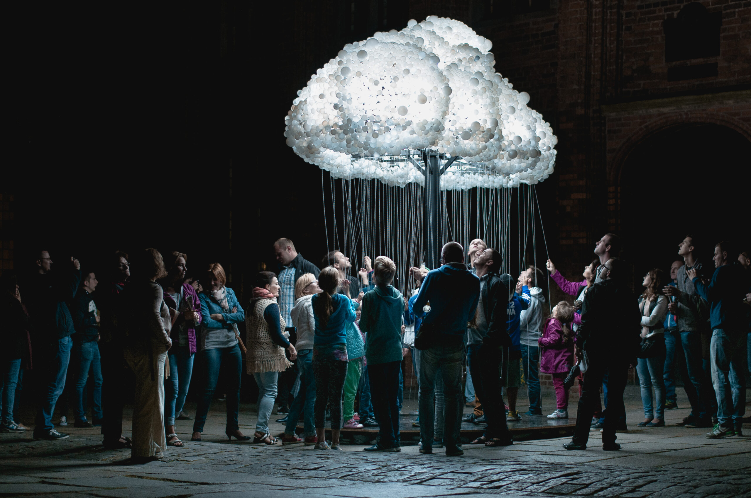 instalacja świetlna przedstawiająca chmurę z wiszącymi sznurkami, za które ciągną ludzie